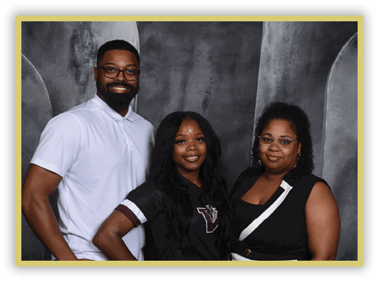 Three adults stand closely together smiling against a dark textured backdrop, with one wearing a white polo shirt, another in a black shirt with a logo, and the third in a black sleeveless top with glasses. Description generated by AI