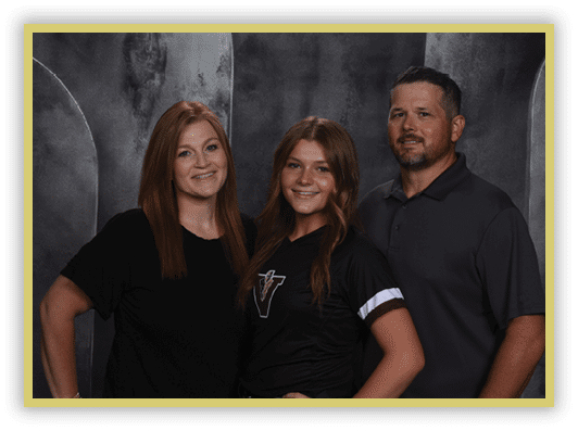 Three adults stand closely together smiling against a dark textured backdrop, with one wearing a black shirt with a logo and the others in solid dark tops. Description generated by AI