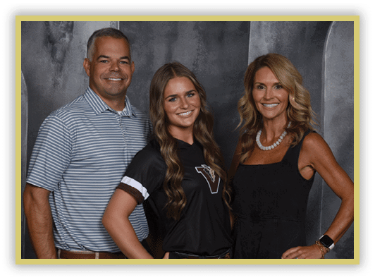 Three adults stand closely together smiling against a dark gray backdrop, with one wearing a striped polo shirt, another in a black sports jersey, and the third in a black sleeveless dress with a necklace. Description generated by AI