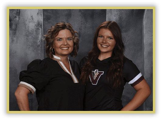 Two adults stand side by side smiling against a gray textured backdrop, one wearing a black blouse with puffed sleeves and the other wearing a black sports jersey with a “V" logo. Description generated by AI