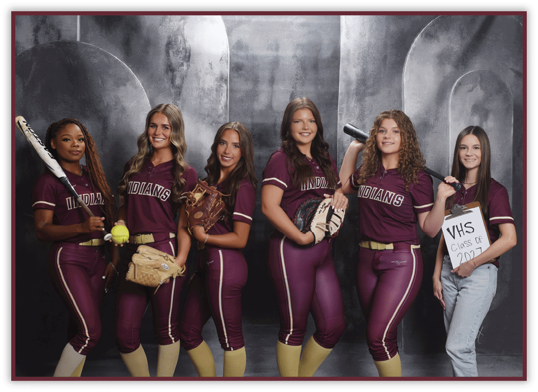 Six people wearing matching maroon softball uniforms and holding softball equipment pose together in front of a gray backdrop, with one person holding a sign that reads “VHS Class of 2024." Description generated by AI