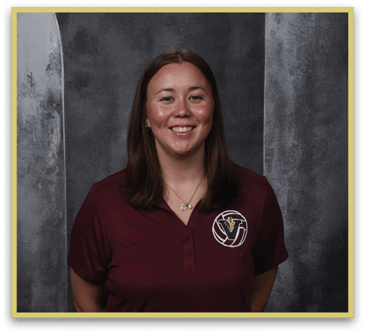 A person with shoulder-length straight hair is smiling and wearing a maroon polo shirt with a logo on the left chest against a gray textured background. Description generated by AI
