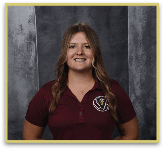 A person with long wavy hair wearing a maroon polo shirt with a logo and a cross necklace is smiling against a dark textured background. Description generated by AI