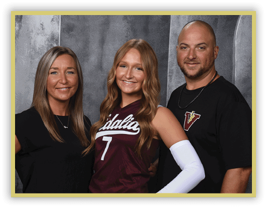Three adults stand closely together smiling, with one wearing a sports jersey and arm sleeve, and the others dressed in casual clothing against a gray backdrop. Description generated by AI