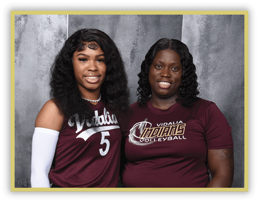 Two people with curly hair wearing maroon Vidalia volleyball shirts smile while posing against a gray backdrop. Description generated by AI