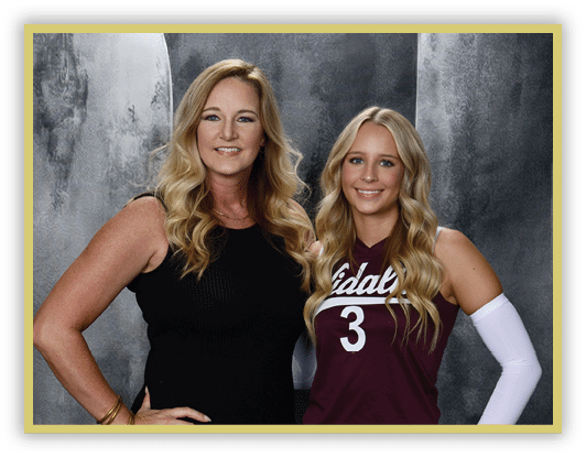 Two adults with long wavy hair stand side by side smiling, one wearing a black sleeveless dress and the other in a maroon sports jersey with the number three and a white arm sleeve, against a gray textured backdrop. Description generated by AI