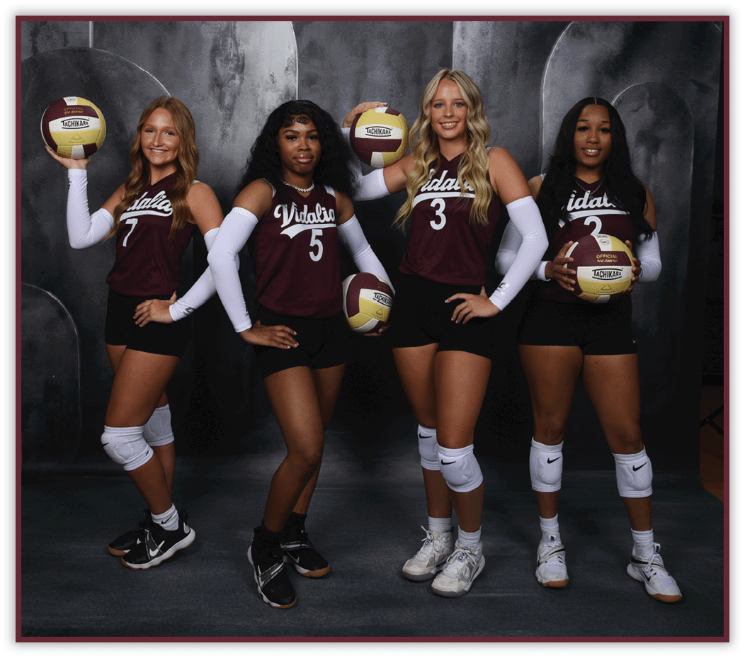 Four adults wearing matching sports uniforms and knee pads pose holding volleyballs against a dark backdrop. Description generated by AI