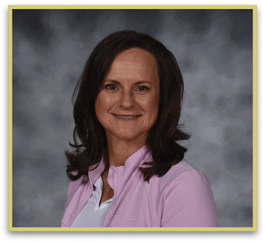 A person with shoulder-length dark hair is smiling and wearing a light pink jacket over a white top against a gray blurred background. Description generated by AI