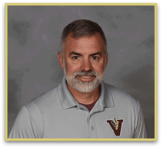 A person with short hair and a beard is smiling and wearing a light gray polo shirt with a maroon and gold “V" logo on the chest against a gray background. Description generated by AI