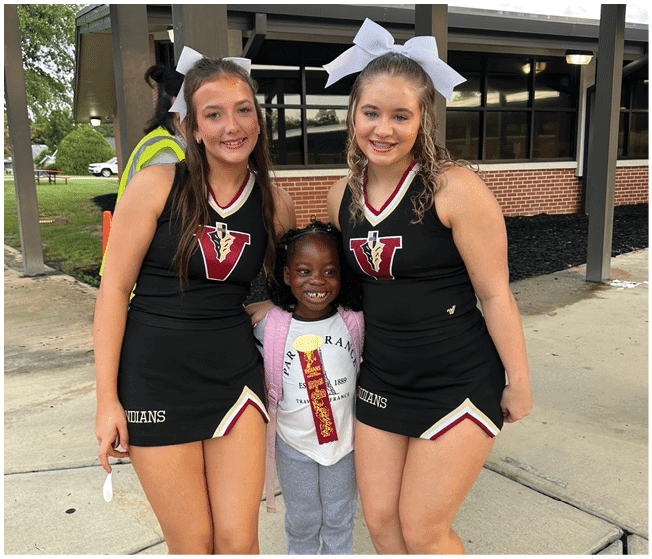 Two adults wearing matching cheerleading uniforms with large white bows in their hair are smiling outdoors near a building. Description generated by AI