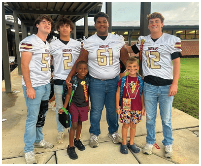 Five people wearing football jerseys stand together under a covered outdoor structure with two children in front smiling. Description generated by AI