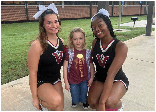Two adults wearing cheerleading uniforms and bows smile while standing on either side of a child holding a small item outdoors near a brick building. Description generated by AI
