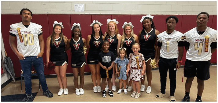 A group of people wearing cheerleading uniforms and sports jerseys stand together indoors against a maroon and beige wall, smiling and posing for the photo. Description generated by AI
