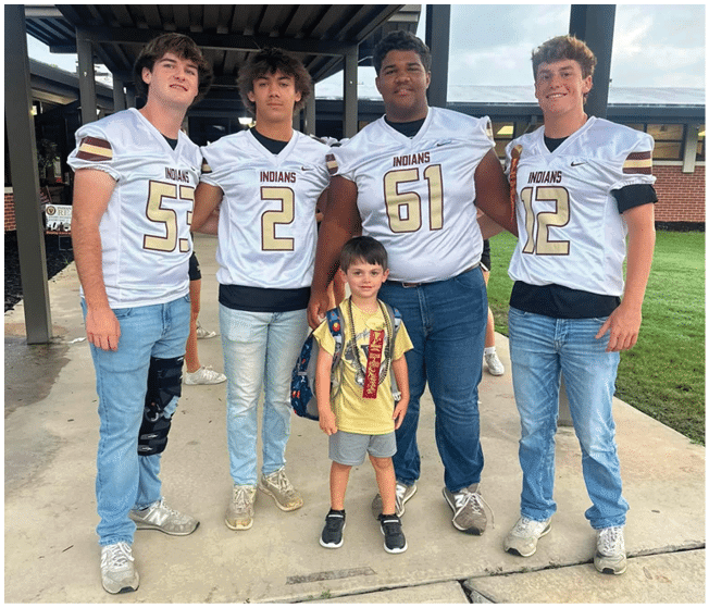 Four people wearing matching sports jerseys stand under a covered outdoor area with a child in front of them. Description generated by AI