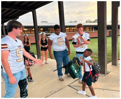 Several people wearing sports jerseys and cheerleading uniforms are gathered outside near a building, with one person holding a backpack and another smiling while interacting with a child carrying a backpack. Description generated by AI