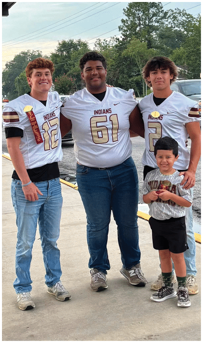 Three people wearing football jerseys stand on a sidewalk with one child in casual clothing in front of them, all smiling and posing for the photo near parked cars and trees. Description generated by AI