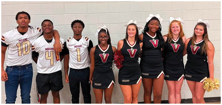Seven people wearing cheerleading uniforms and three people wearing sports jerseys stand side by side against a gymnasium wall, smiling and posing for the photo. Description generated by AI