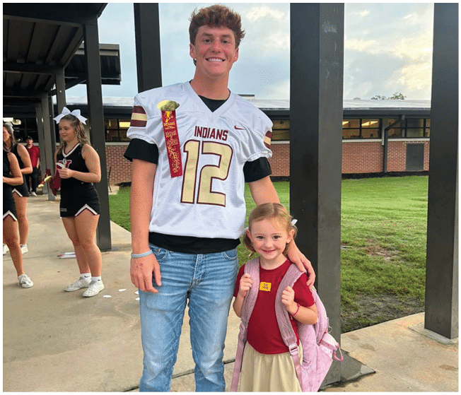 An adult wearing a sports jersey with the number 12 and a ribbon stands next to a smiling child with a backpack giving thumbs up outside near a building with cheerleaders in the background. Description generated by AI