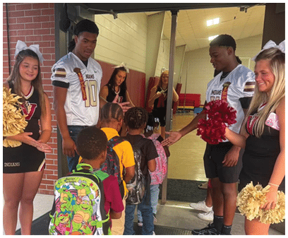People in cheerleading uniforms and sports jerseys greet children with backpacks as they enter a school hallway. Description generated by AI