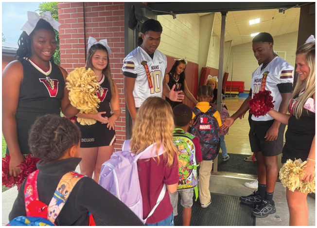 Several people wearing cheerleading and sports uniforms are interacting with children carrying backpacks near a brick building entrance. Description generated by AI