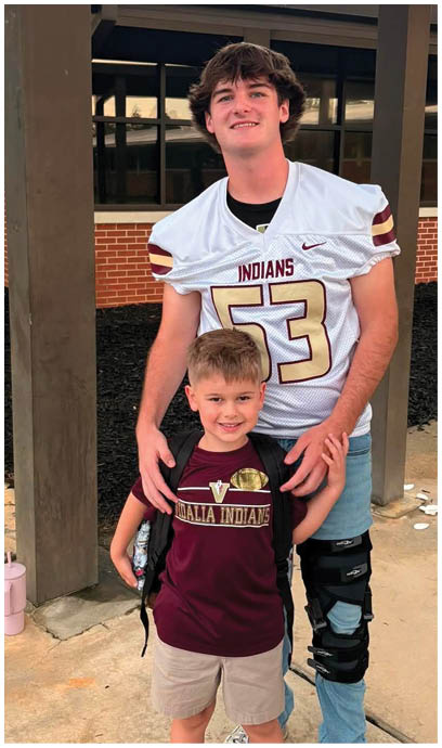 A person wearing a football jersey with the number 53 and a knee brace stands behind a smiling child wearing a maroon sports shirt with a logo, both posing outdoors near a building. Description generated by AI