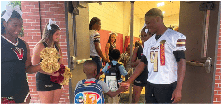 People wearing sports and cheerleading uniforms are gathered near a doorway, engaging in conversation and holding pom-poms. Description generated by AI