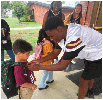 An adult wearing a sports jersey is helping a child with a maroon garment while another child with a pink backpack stands nearby outside on a sidewalk. Description generated by AI