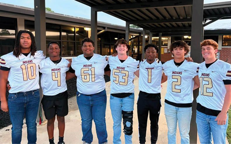 Seven people wearing matching sports jerseys with the word “Indians" stand side by side outdoors under a covered walkway, smiling and posing for the photo. Description generated by AI