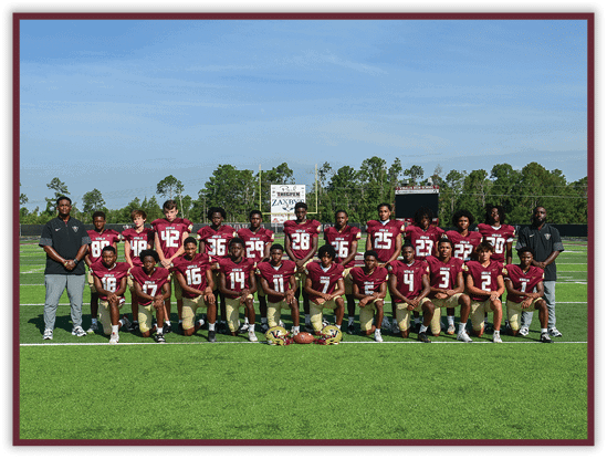 A group of people wearing maroon and gold football uniforms pose for a team photo on a green football field with two coaches standing on either side. Description generated by AI