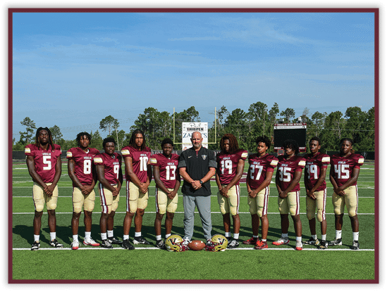 A group of people wearing maroon and gold football uniforms stand on a football field with a coach in the center holding a football. Description generated by AI