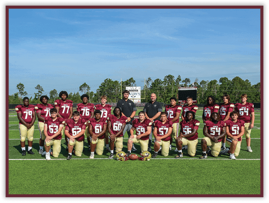 A group of people wearing maroon and gold football uniforms pose together on a green field with a clear blue sky in the background. Description generated by AI