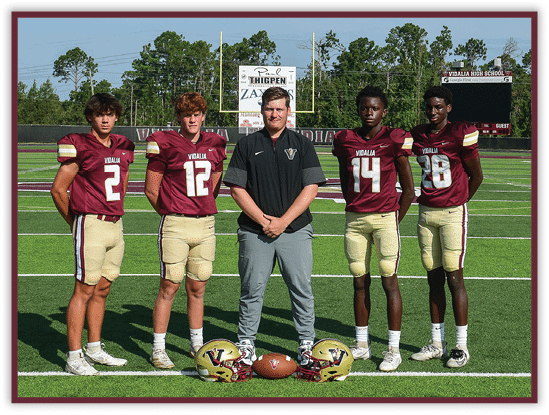 Five people wearing football uniforms and one person in casual sportswear stand on a football field with helmets and a football placed on the ground in front of them. Description generated by AI