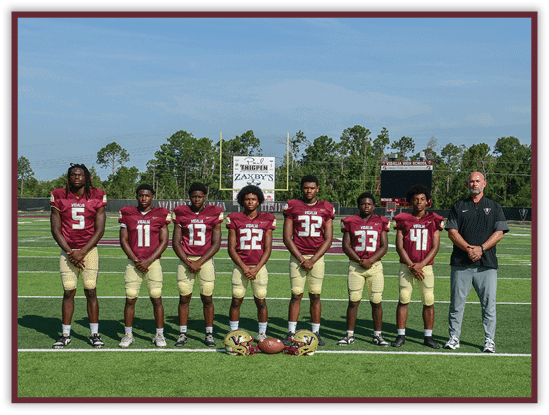 Seven people wearing matching football uniforms stand in a line on a football field with helmets and a football placed on the ground in front of them and a scoreboard visible in the background. Description generated by AI