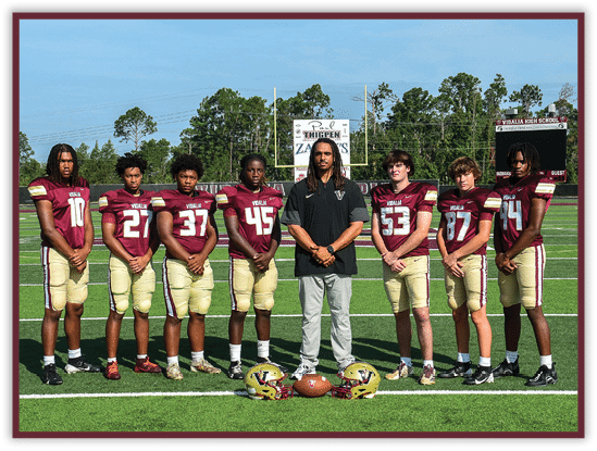 Seven people wearing matching football uniforms stand on a football field with one person in casual clothing standing in the center, and two football helmets and a football placed on the ground in front of them. Description generated by AI