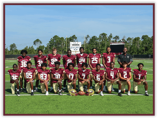 A group of people wearing maroon and gold football uniforms pose together on a football field with trees and a scoreboard in the background. Description generated by AI