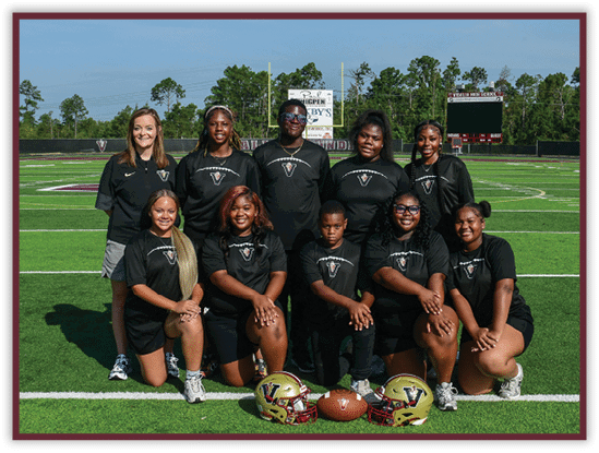 A group of adults and one child wearing matching black sports shirts pose on a football field with two helmets and a football placed in front of them. Description generated by AI