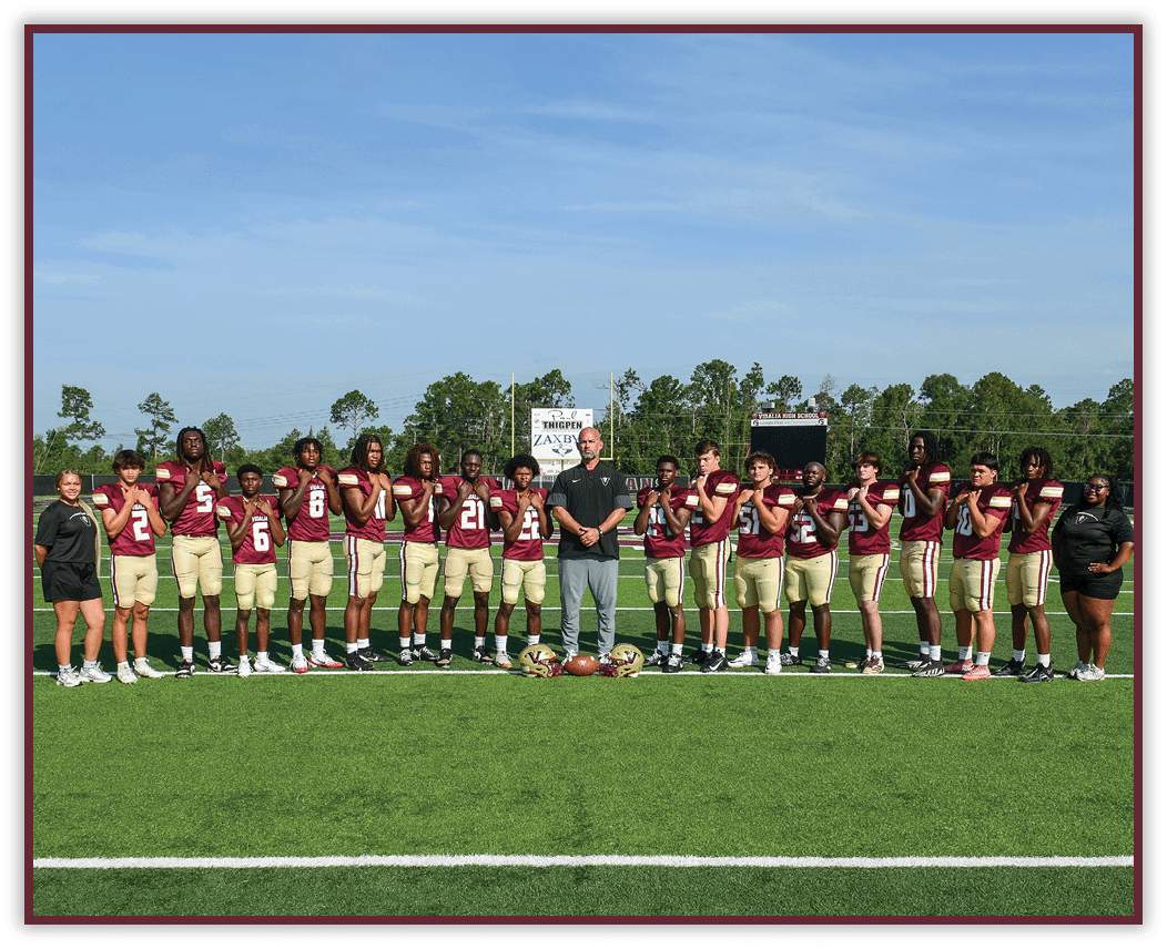 A group of people wearing matching sports uniforms and two adults stand in a line on a football field with helmets and a football placed on the ground in front of them. Description generated by AI