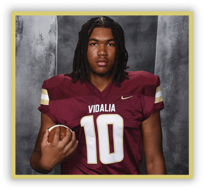 A person with shoulder-length dreadlocks wearing a maroon football jersey with the number 10 and the word Vidalia is holding a football. Description generated by AI
