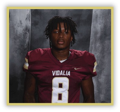 A person with short dreadlocks wearing a maroon football jersey with the word Vidalia and the number 8 stands against a dark gray backdrop. Description generated by AI