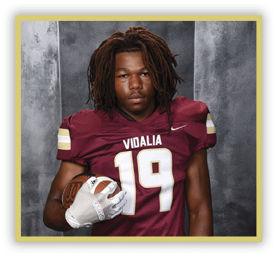 A person with dreadlocks wearing a maroon football jersey with the number 19 and the word Vidalia is holding a football against a gray backdrop. Description generated by AI
