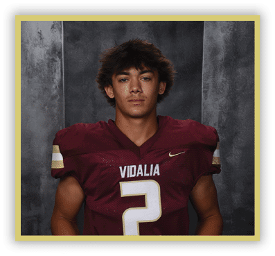 A person with medium-length tousled hair wearing a maroon football jersey with the word Vidalia and the number 2 stands against a dark textured background. Description generated by AI