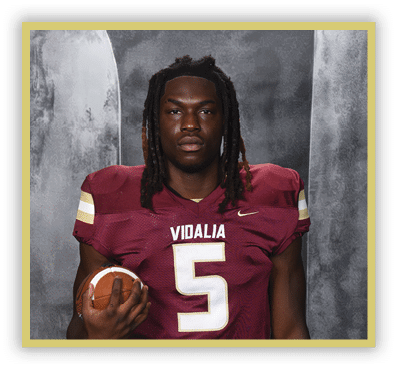 A person with long dreadlocks wearing a maroon football jersey with the number five and the word Vidalia is holding a football against a gray textured background. Description generated by AI