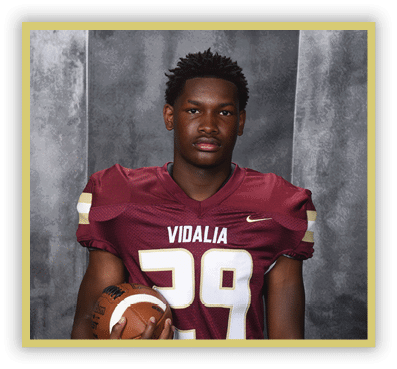 A person wearing a maroon football jersey with the number 29 and the word Vidalia is holding a football against a gray backdrop. Description generated by AI