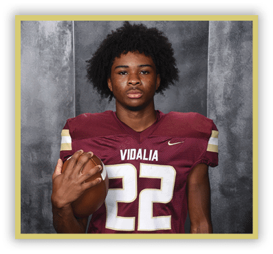 Person with curly hair wearing a maroon football jersey with the number 22 and holding a football against a gray backdrop. Description generated by AI