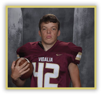 A person wearing a maroon football jersey with the number 42 and the word Vidalia is holding a football against a dark gray background. Description generated by AI