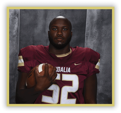 A person wearing a maroon football jersey with the word Vidalia and the number 5 stands against a gray textured backdrop. Description generated by AI