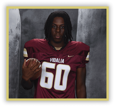 A person with shoulder-length dreadlocks wearing a maroon football jersey with the number 60 and the word Vidalia is holding a football against a gray backdrop. Description generated by AI