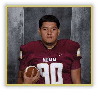 A person wearing a maroon football jersey with the number 90 and the word Vidalia is holding a football against a gray backdrop. Description generated by AI