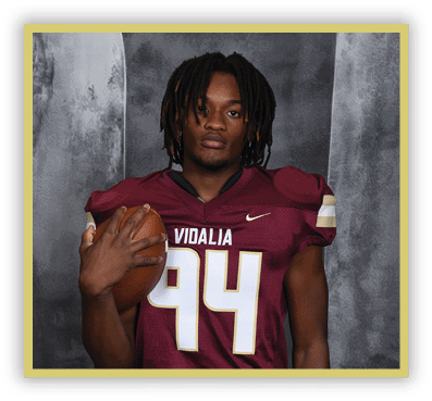Person with dreadlocks wearing a maroon football jersey holding a football against a gray textured background. Description generated by AI