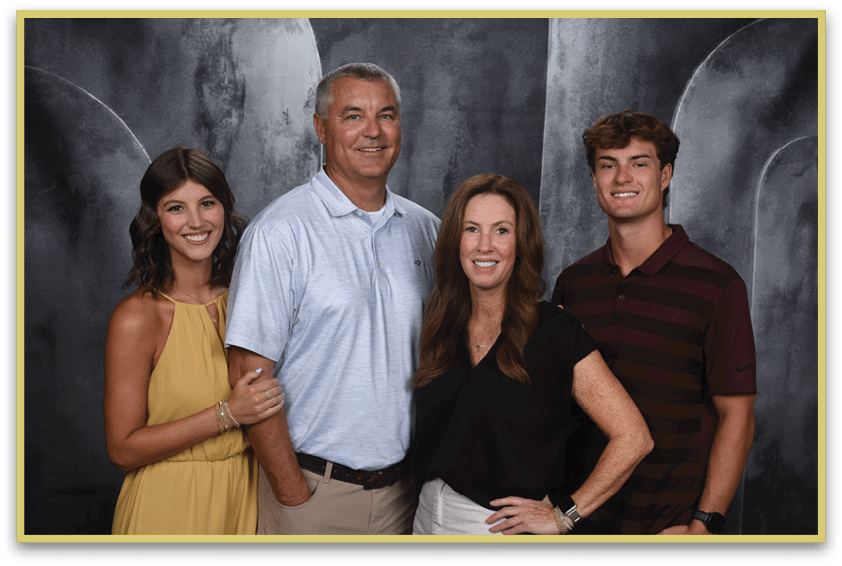Four adults stand closely together smiling against a dark textured backdrop, with one wearing a yellow dress, one in a light blue polo shirt, one in a black top, and one in a dark striped polo shirt. Description generated by AI